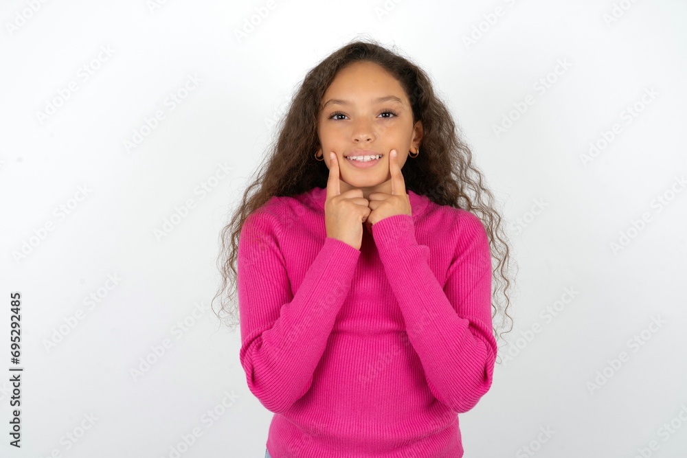 Happy Beautiful teen girl wearing pink sweater over white background with toothy smile, keeps index fingers near mouth, fingers pointing and forcing cheerful smile Happy Beautiful teen girl wearing pink sweater over white background with toothy smile, keeps index fingers near mouth, fingers pointing and forcing cheerful smile