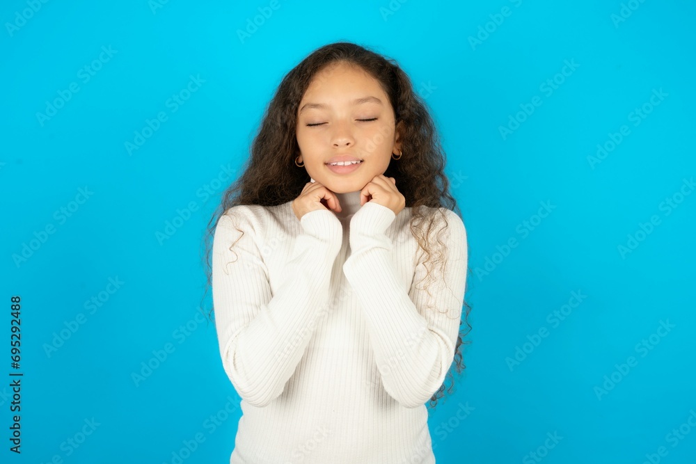 Young beautiful teen girl wearing white turtleneck over blue background grins joyfully, imagines something pleasant, copy space. Pleasant emotions concept. Young beautiful teen girl wearing white turtleneck over blue background grins joyfully, imagines something pleasant, copy space. Pleasant emotions concept.