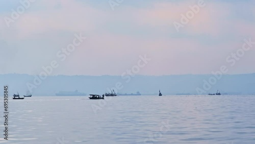 Blurred small wooden motorboats carrying people sail and sway with the waves past freight ferries, large tankers emitting pollution fumes, and fishing boats nearby