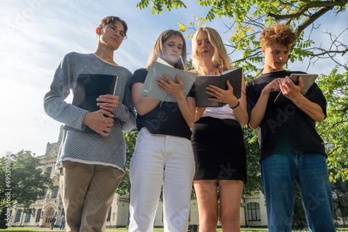 Focused young students take notes while standing in front of campus. Group of students listens to professor taking notes