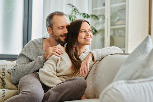 smiling man with closed eyes embracing wife on comfortable couch in living room, child-free couple