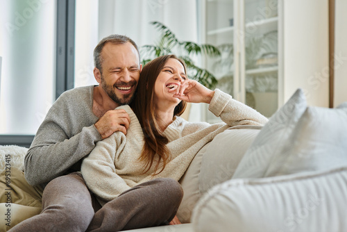 laughing man with closed eyes embracing wife on comfortable couch in living room, child-free couple