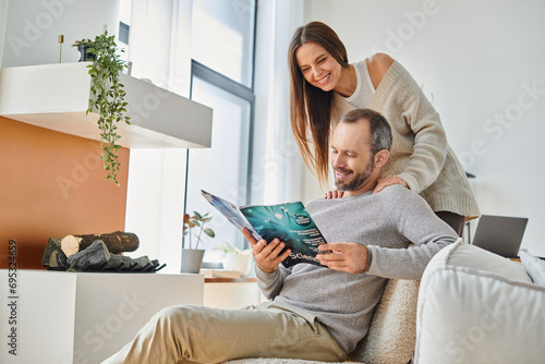 joyful man reading science magazine near smiling wife on couch in living room, child-free couple