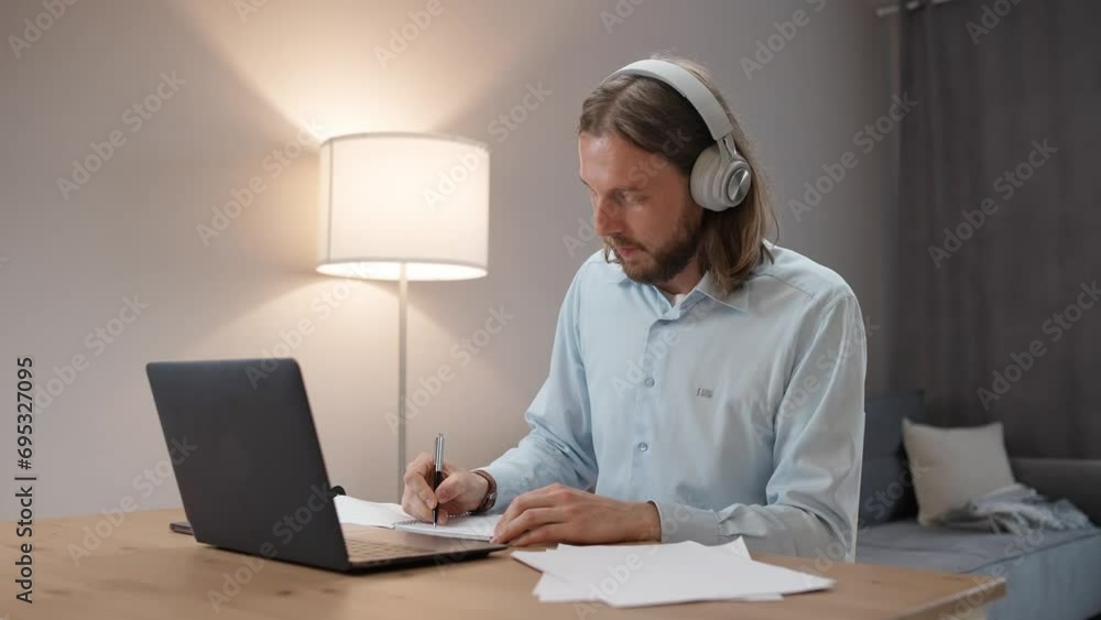 Freelancer at remote work, man in headphones in a online meeting on a video conference, workplace in living room, person at a remote job, man writes in a notebook. Freelancer at remote work, man in headphones in a online meeting on a video conference, workplace in living room, person at a remote job, man writes in a notebook.