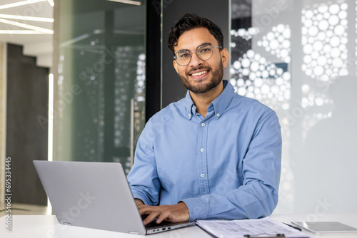 Portrait of young successful businessman inside office, satisfied joyful Indian man looking at camera, worker at workplace using laptop, typing on keyboard, financier accountant in shirt photo
