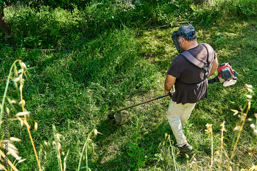 Gardener mowing weeds in the garden with string grass trimmer. Man using lawnmower in yard, cutting green grass.