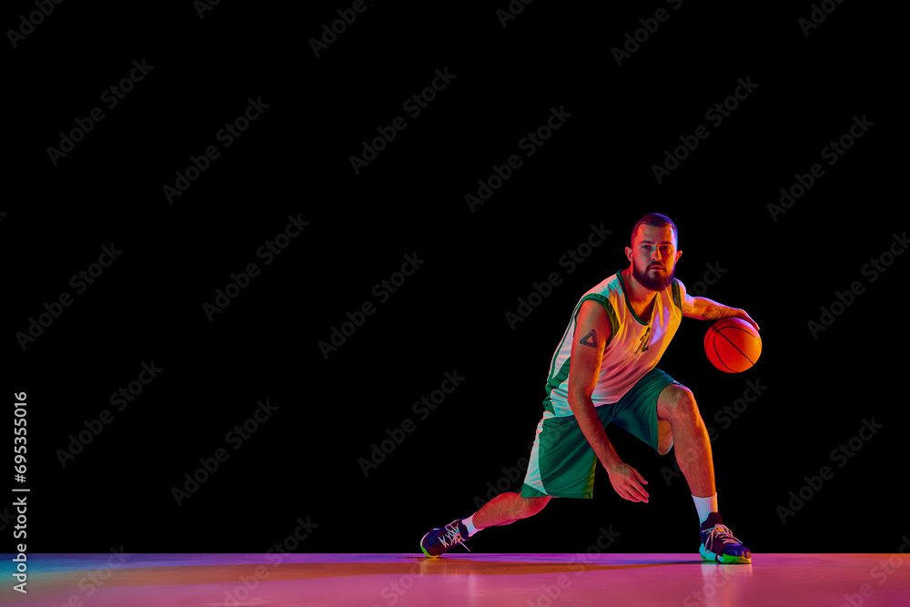Dynamic portrait of professional basketball player honing skills with precision dribbling against black background in neon light.