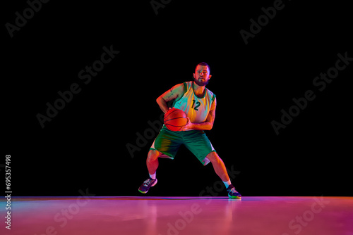 Full length portrait of athletic man, professional basketball player actively training before match against black background in neon light.