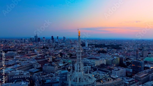 Aerial view of the statue of the Madonna on the central spire of the Duomo and the city s cathedral at dawn. Roofs of houses and skyscrapers. Sun over the horizon. duomo square. Italy Milan 16.11.2023