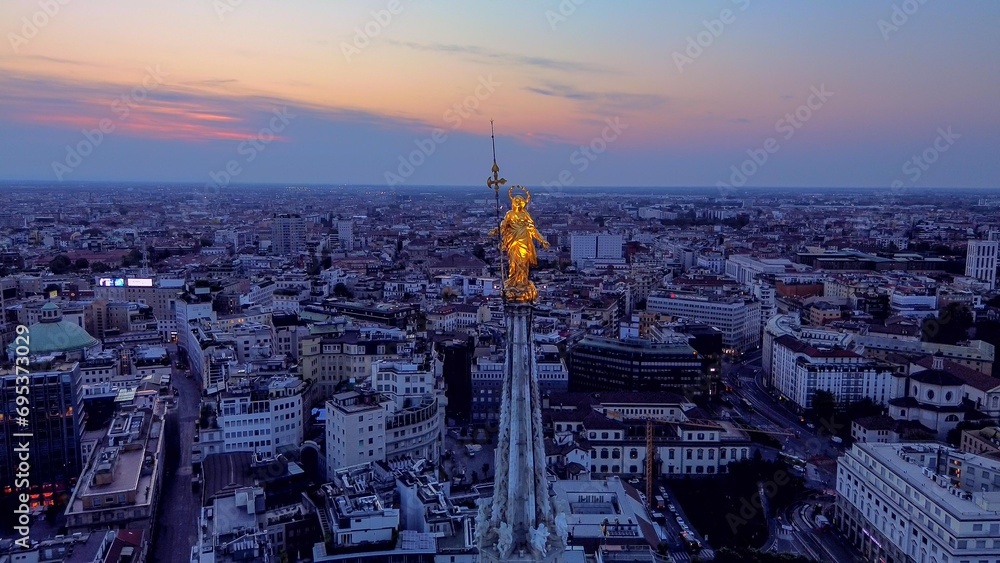 Aerial view of the statue of the Madonna on the central spire of the Duomo and the city's cathedral at dawn. Roofs of houses and skyscrapers. Sun over the horizon. duomo square. Italy Milan 16.11.2023