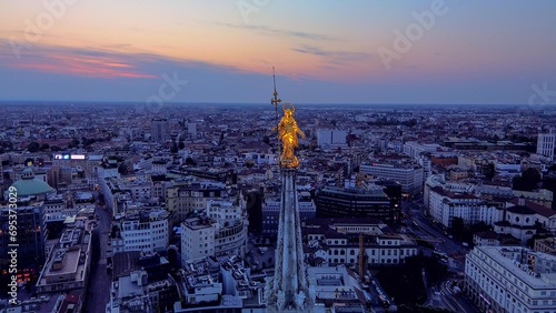 Aerial view of the statue of the Madonna on the central spire of the Duomo and the city s cathedral at dawn. Roofs of houses and skyscrapers. Sun over the horizon. duomo square. Italy Milan 16.11.2023