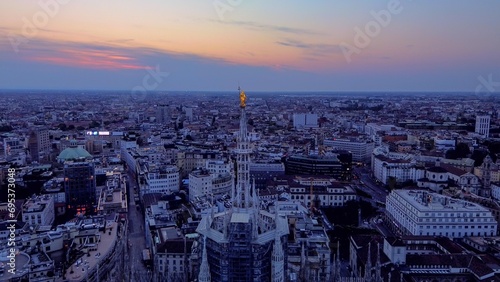Aerial view of the statue of the Madonna on the central spire of the Duomo and the city's cathedral at dawn. Roofs of houses and skyscrapers. Sun over the horizon. duomo square. Italy Milan 16.11.2023 © VILTVART