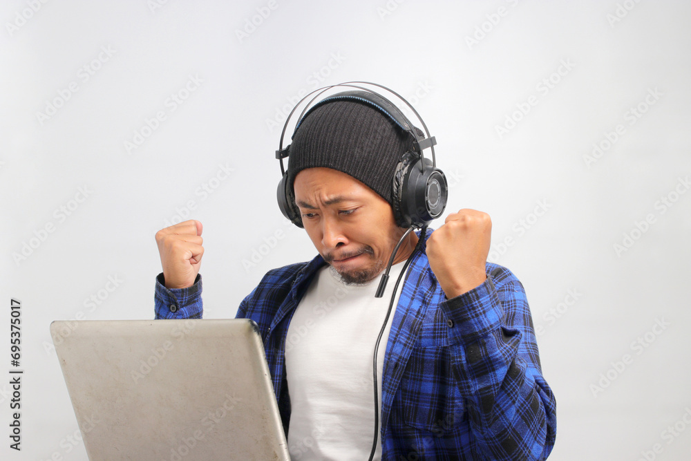 Frustrated Asian man, donning a beanie hat, headphones, and a casual shirt, raises his clenched fists while working remotely from home with a laptop, Feels annoyed, and expresses anger through yelling Frustrated Asian man, donning a beanie hat, headphones, and a casual shirt, raises his clenched fists while working remotely from home with a laptop, Feels annoyed, and expresses anger through yelling