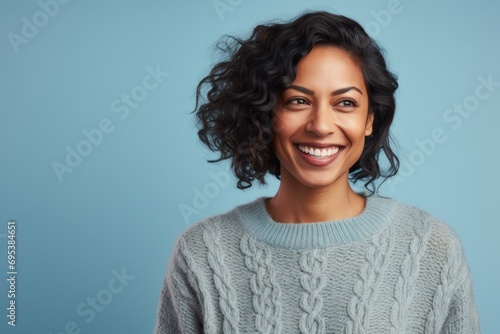 Portrait of a grinning indian woman in her 30s dressed in a warm wool sweater against a pastel blue background. AI Generation © Markus Schröder
