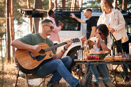 Man is playing guitar, by table. Group of friends are together in the forest