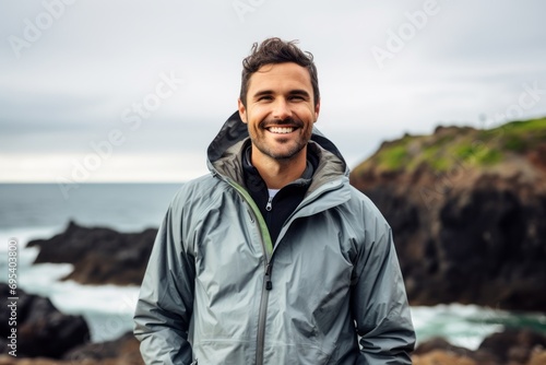 Portrait of a smiling man in his 30s wearing a functional windbreaker against a rocky shoreline background. AI Generation