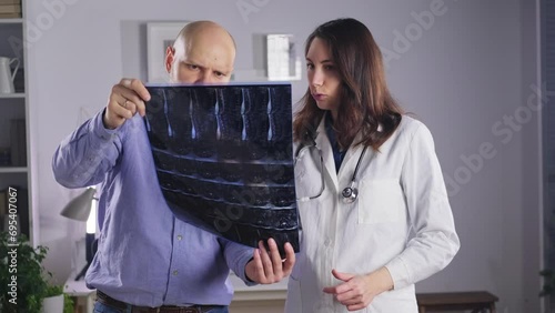 A female doctor in a white coat holds an X-ray of the bones of the spine in her hands and shows the patient his injuries. Medicine and treatment of protrusion and fractures