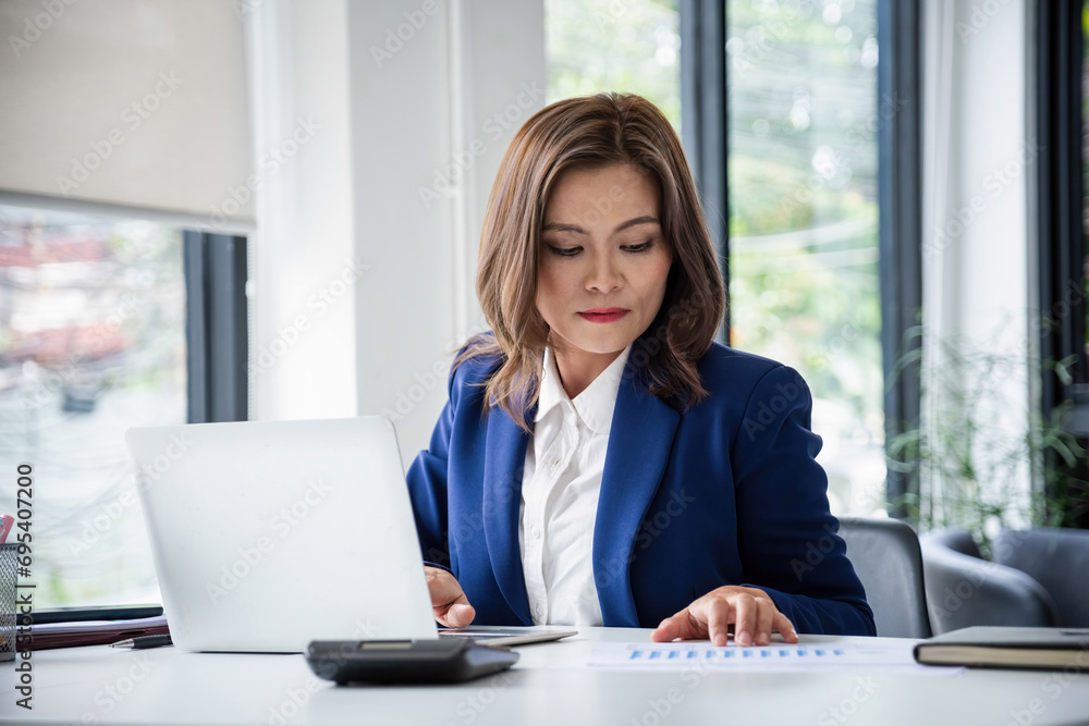 Businesswoman sitting at desk on couch in workplace or at home working on laptop and analyzing data on charts and graphs and writing on papers Businesswoman sitting at desk on couch in workplace or at home working on laptop and analyzing data on charts and graphs and writing on papers
