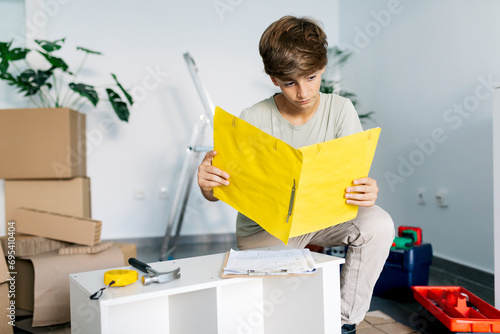 Focused boy kneeling and reading notes on yellow clipboard photo