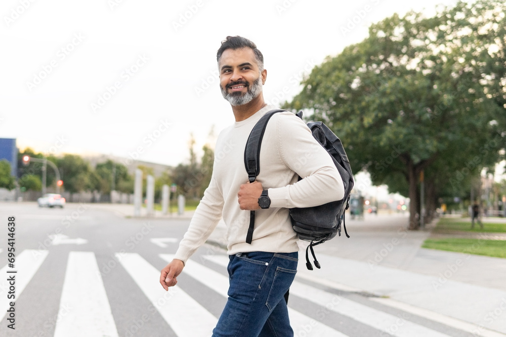 Happy Indian man in casual clothing with backpack looking away while crossing the street Happy Indian man in casual clothing with backpack looking away while crossing the street