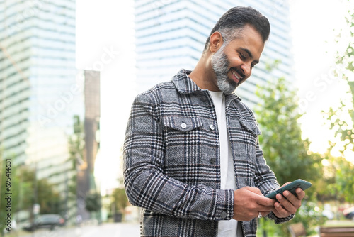 Bearded Indian businessman using cellphone and smiling while standing outside photo