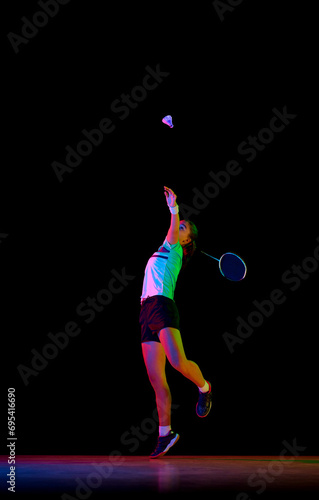 Portrait of young girl, skilled badminton player practicing with intensity against black background in neon light. Concept of sport, active, healthy lifestyle, strength and power, action. Copy space.