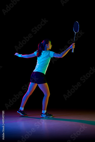 Full length rear view portrait of young girl, skilled badminton player practicing with intensity against black background in neon light. Concept of sport, active lifestyle, action. Copy space.