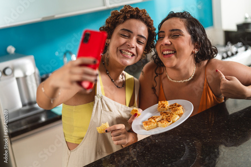 Amigas tirando uma selfie na cozinha, segurando o prato que prepararam juntas.
