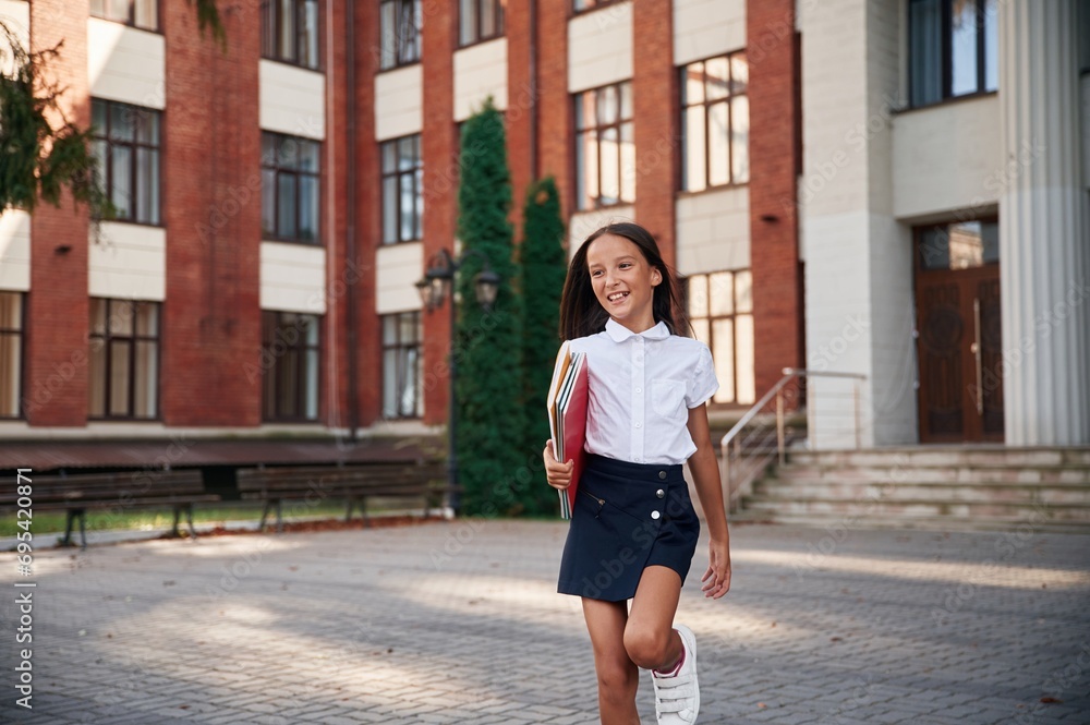 Standing, front view, holding books. School girl in uniform is outdoors near the building Standing, front view, holding books. School girl in uniform is outdoors near the building