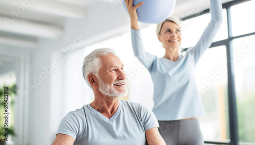 Senior citizens engaging in physical exercise, a man with a gym ball and a woman stretching against the backdrop of a bright room. The concept of activity of the elderly