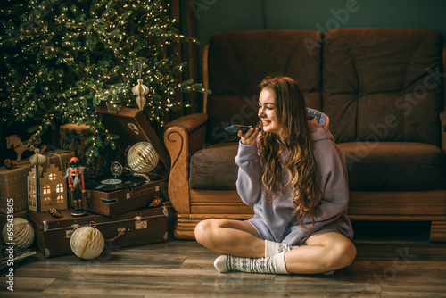 Young woman with headphones listens to music from a vinyl record, sitting on floor near a glowing Christmas tree, girl with smartphone. Records a voice message