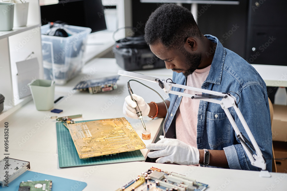 Side view portrait of African American man fixing circuit board with soldering iron in computer repair workshop, copy space