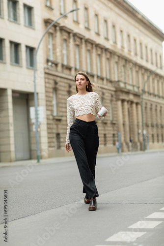 young woman in lace top and high waist pants holding coffee to go on street in Berlin, Germany
