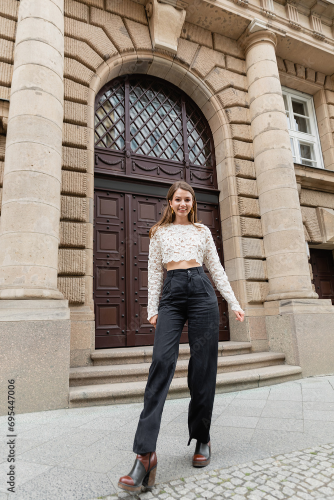 cheerful young woman in lace top and high waist pants looking at camera on urban street in Berlin