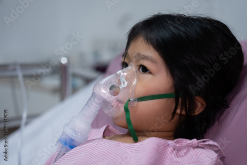 A little Asian girl has an oxygen mask and breathing through a nebulizer at the hospital. Concept of bronchitis, respiratory and Medical treatment, inhaling medicine, mist, from a nebulizer.