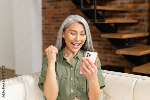 Surprised senior woman looking at the phone screen sitting on the coach at home, an wondered elderly lady saw unexpected good news or received pleasantly message, sales proposal on the smartphone