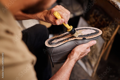 Hand of unrecognizable male shoemaker spreading glue over leather sole of grey boot while sitting in workshop and creating new footwear photo