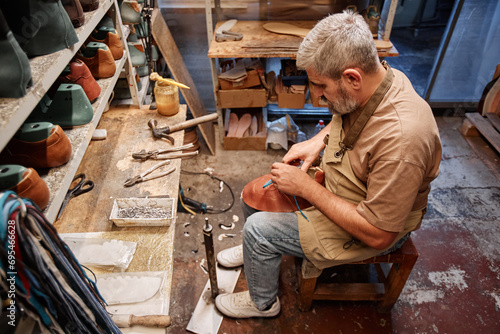 High angle of mature experienced shoemaking master in casualwear sitting in front of table and shelves with footwear workpieces photo