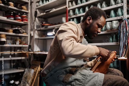 Young African American male owner of shoemaking workshop fixing upper part of unfinished boot to sole while sitting by workplace photo