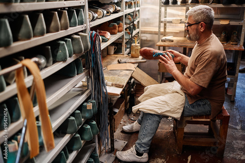 Side view of mature professional shoemaker holding brown boot workpiece while sitting by table with long shelves above
