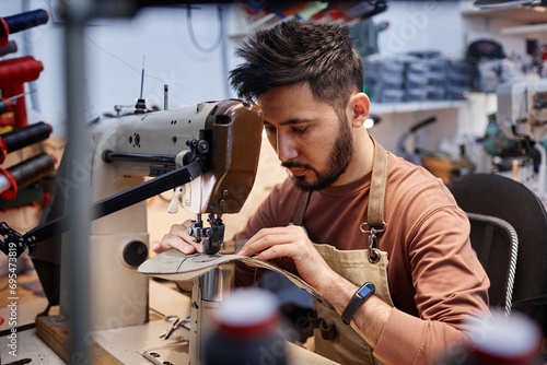 Young Asian man in workwear sitting in front of sewing machine in workshop and putting together details of new footwear