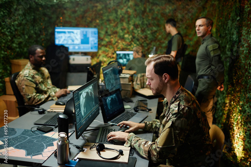 Side view of young male military officer typing on keyboard and looking at computer screen with graphic data among other specialists