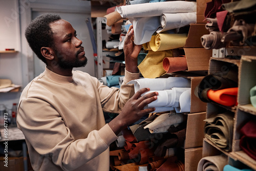 Young African American male leatherworker choosing suitable color of suede or leather textile for creating new items for sale in craft shop photo