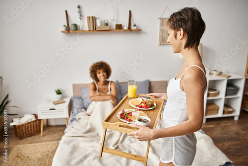 happy woman holding tray with pancakes and glass of orange juice near girlfriend, breakfast in bed