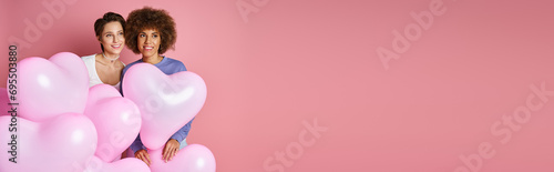 Valentines day banner, dreamy interracial lesbian couple smiling near heart shaped balloons © LIGHTFIELD STUDIOS