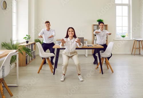 Full length portrait of young business people doing stretching sit-ups exercises standing at workplace in office. Group of smiling company employees exercising for health during a break from a work.
