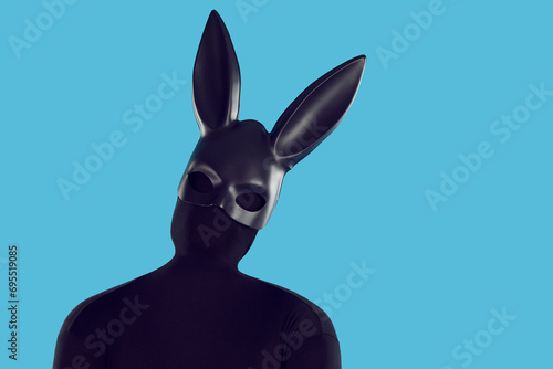 Studio portrait fashion model disguised in rabbit costume. Anonymous adult man in faceless monochromatic black bodysuit and long eared bunny mask tilts head isolated like silhouette on blue background photo