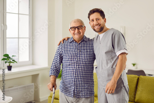 Health care worker with an elderly man smiling, hugging and looking at the camera.Young cheerful nurse helping a senior patient to walk with his crutches. Physiotherapist helping old man in rehab.
