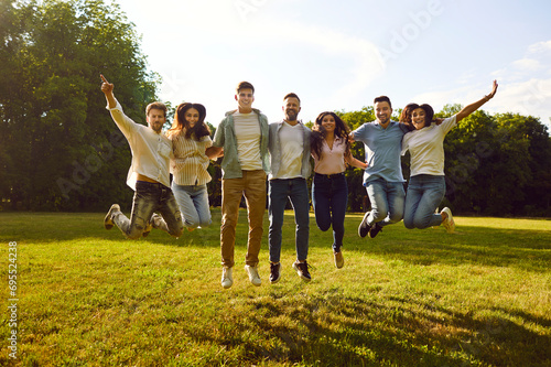 Group of cheerful young people jumping high together having fun in park on summer sunny day. Funny friends in casual clothes hugging in row and doing jump together. Concept of friendship and summer.