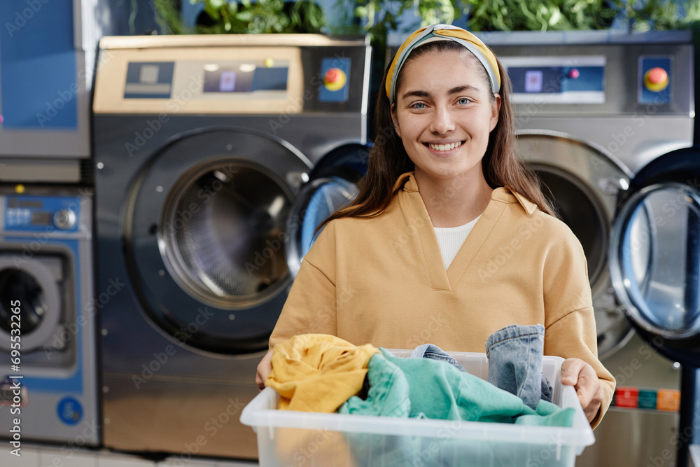 Pretty girl in casualwear holding plastic container with clean clothes against washing machines in laundry and looking at camera Pretty girl in casualwear holding plastic container with clean clothes against washing machines in laundry and looking at camera